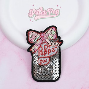 Pode incluir: Um patch de lantejoulas em forma de frasco com um laço no topo. O patch tem uma borda preta, lantejoulas prateadas e as palavras "Patch Pop" e "Dior" em rosa e vermelho. O laço é rosa e iridescente.