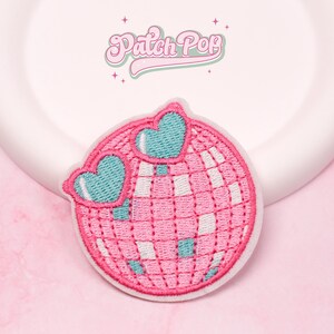 Puede incluir: Parche bordado con una bola de discoteca rosa con gafas de sol en forma de corazón turquesa. El parche tiene un borde blanco y el texto "Patch Pop" en cursiva encima de la bola de discoteca. El fondo es rosa claro.