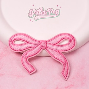 Puede incluir: Un parche bordado rosa con forma de lazo con un diseño detallado. El parche tiene forma de lazo con un nudo central y extremos en bucle. El texto "Patch Pop" está encima del lazo.