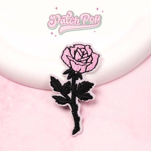 Könnte beinhalten: Ein rosa Rosenaufnäher mit schwarzem Stiel und Blättern. Der Aufnäher hat einen weißen Rand und ist vor einem rosa Hintergrund platziert. Der Text "Patch Pop" steht in einer Retro-Schriftart über der Rose.