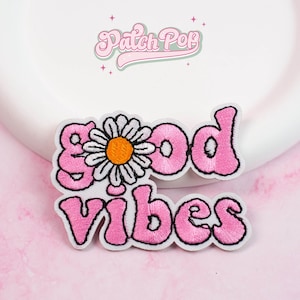 Puede incluir: Parche bordado con las palabras "Good Vibes" en letras rosas con burbujas. Una margarita blanca con un centro naranja reemplaza la letra "o" en "Good". El parche tiene un borde blanco y el texto "Patch Pop" en una fuente retro.