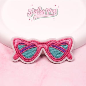 Peut inclure: Écusson brodé représentant des lunettes de soleil en forme de cœur avec des montures roses, des verres turquoise et un accent violet. L'écusson a une bordure blanche et le texte "Patch Pop" en cursive.