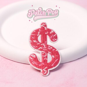 May include: A red dollar sign patch with white trim. The patch is embroidered with a textured pattern and sits on a white surface. The text "Patch Pop" is above the patch in a retro font.
