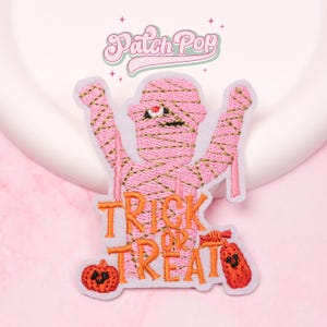 May include: A pink embroidered patch featuring a mummy with outstretched arms. The mummy has gold stitching and a single red eye. The words "TRICK OR TREAT" are in orange letters below the mummy, with a pumpkin and a candy bag on either side. The patch has the text "Patch Pop" at the top.