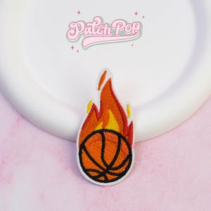May include: Embroidered patch featuring a basketball engulfed in flames. The basketball is orange with black lines, and the flames are yellow and red. The patch has a white border and the text "Patch Pop" in pink.