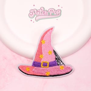 May include: Embroidered pink witch hat patch with yellow stars, a purple band, and a gold buckle. A black spiderweb design is on the side. The patch has a white border and the text "Patch Pop" in a cursive font.