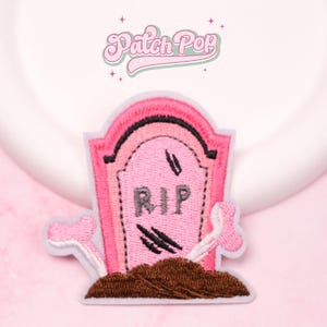 May include: Embroidered patch featuring a pink tombstone with the letters "RIP" in gray. The patch includes brown soil, a pair of crossed bones, and the text "Patch Pop" in a cursive font. The design is set against a pink background.