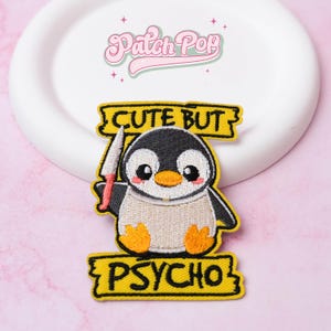 Op de afbeelding: Geborduurde patch met een cartoonpinguïn met een mes. De pinguïn is zwart, wit en bruin, met oranje voeten en een roze blos. De patch heeft de tekst "CUTE BUT PSYCHO" in het geel.