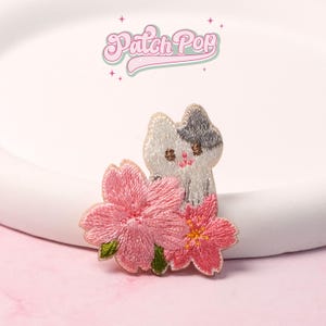 May include: Embroidered patch featuring a white and gray cat with pink cherry blossoms. The patch has the text "Patch Pop" in a pink, cursive font. The cat has small, embroidered details and is set against a white background.