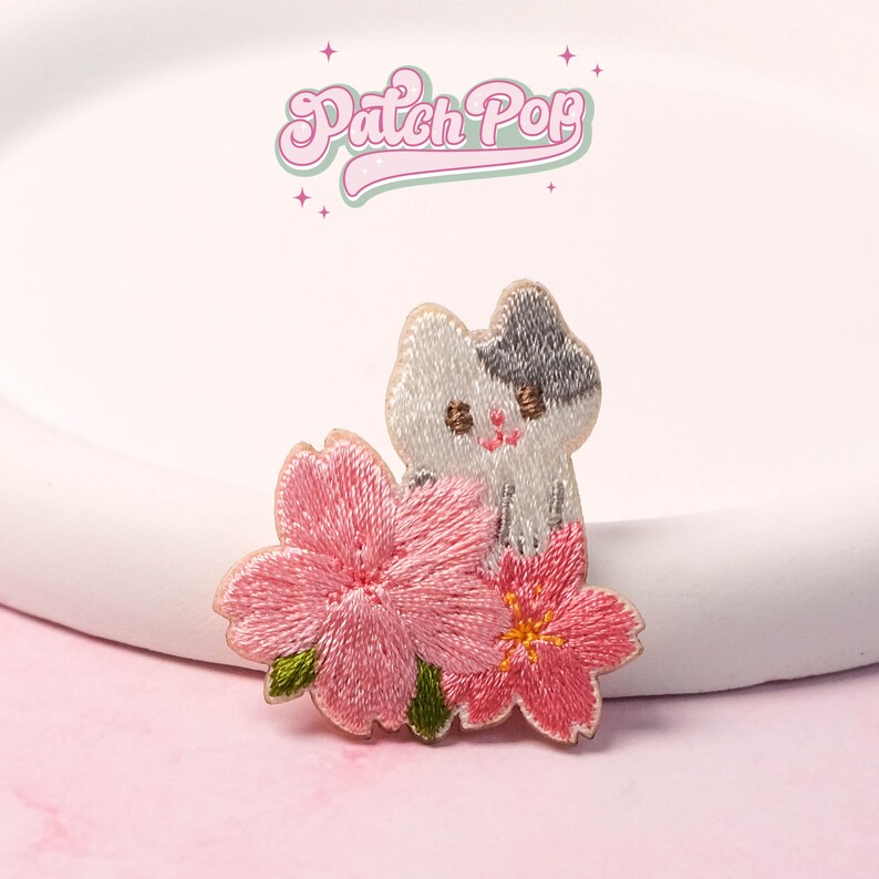 May include: Embroidered patch featuring a white and gray cat with pink cherry blossoms. The patch has the text "Patch Pop" in a pink, cursive font. The cat has small, embroidered details and is set against a white background.
