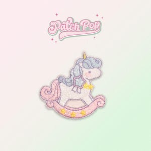 Puede incluir: Un parche bordado que representa un unicornio rosa y morado montando un caballo de balancín. El unicornio tiene una crin y una cola azules, y el caballo de balancín está decorado con estrellas amarillas. El parche dice "Patch Pop" en la parte superior.
