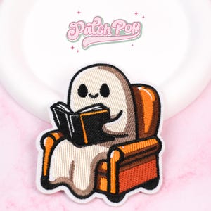 Op de afbeelding: Geborduurde patch met een vriendelijk spook dat een boek leest in een bruine en oranje fauteuil. De patch heeft een witte rand en de tekst "Patch Pop" in roze boven het spook.