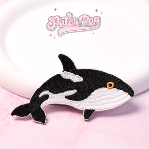 Puede incluir: Parche bordado con un diseño de orca en blanco y negro. El parche tiene un ojo naranja y detalles blancos en el vientre y las aletas. El texto "Patch Pop" está en rosa, en una fuente retro.