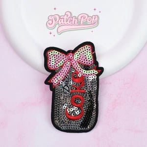 Può includere: Una toppa di paillettes a forma di lattina di Coca-Cola con un fiocco rosa in cima. La toppa ha un bordo nero e la scritta "COKE" in rosso. Lo sfondo è bianco e rosa, con la scritta "Patch Pop" in alto.