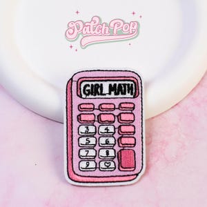 Puede incluir: Un parche bordado rectangular con forma de calculadora, con marco rosa y botones blancos. La pantalla muestra el texto negro GIRL MATH. Los botones presentan números negros y un símbolo de corazón. El parche tiene un borde blanco.