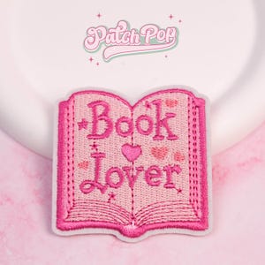 May include: A pink embroidered patch in the shape of an open book. The patch features the words "Book Lover" in a script font, with heart and star accents. The patch has a white border and is set against a soft pink background.