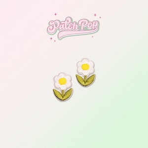 Puede incluir: Dos parches de hierro de margarita blanca con centros amarillos y tallos verdes. Los parches están sobre un fondo rosa claro y verde.