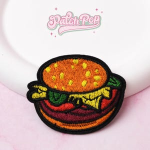 Może przedstawiać: Haftowana naszywka z kolorowym wzorem burgera. Naszywka zawiera bułkę sezamową, sałatę, pomidora i kotleta. Naszywka ma czarną obwódkę i napis "Patch Pop" w kolorze różowym.