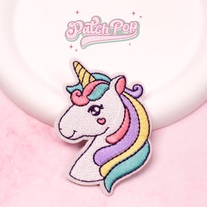 Puede incluir: Parche bordado de unicornio con cuerpo blanco, cuerno amarillo y melena colorida en tonos rosa, morado y turquesa. El parche tiene un detalle en forma de corazón en la mejilla. El texto "Patch Pop" está encima del unicornio.