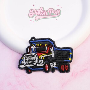May include: Embroidered patch depicting a cartoon dump truck. The truck is mainly black, with red, yellow, and blue details. The patch includes the text "Patch Pop" in a pink, cursive font.