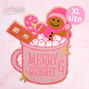 May include: Pink and gold XL size patch featuring a gingerbread man in a pink mug filled with marshmallows. The mug has the words "MERRY & BRIGHT" in pink. A candy cane and chocolate bar are also in the mug. The patch has a gold glitter outline.
