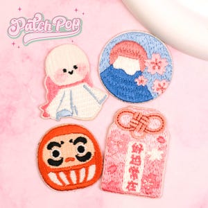 Puede incluir: Cuatro parches bordados sobre una superficie rosa. Los parches incluyen una figura blanca con forma de muñeca con detalles en rosa, un círculo azul con una montaña y flores rosas, una muñeca Daruma roja y un amuleto rosa con texto japonés. El texto "Patch Pop" está en la esquina superior izquierda.