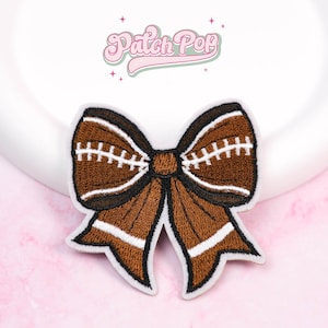 Puede incluir: Parche bordado con forma de lazo, diseñado para parecerse a un balón de fútbol americano. El lazo es marrón con costuras blancas y detalles negros. El parche tiene un borde blanco y el texto "Patch Pop" en rosa.