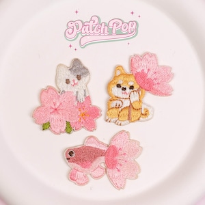 Puede incluir: Parches bordados con un gato, un perro Shiba Inu y un pez dorado, cada uno con flores de cerezo rosas. Los parches son de varios colores, incluyendo blanco, gris, amarillo y rosa. El texto "Patch Pop" está en la parte superior.
