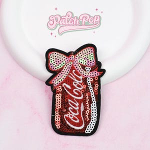 Könnte beinhalten: Ein roter Pailletten-Coca-Cola-Dosen-Aufnäher mit einer Pailletten-Schleife oben. Der Aufnäher hat einen schwarzen Rand und das Coca-Cola-Logo in Weiß. Der Hintergrund ist hellrosa mit dem Text "Patch Pop" oben.