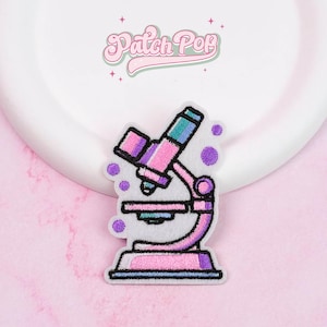 Puede incluir: Parche bordado colorido con un diseño de microscopio. El parche tiene un fondo blanco con un microscopio rosa, morado y turquesa. El texto "Patch Pop" está en rosa encima del microscio.