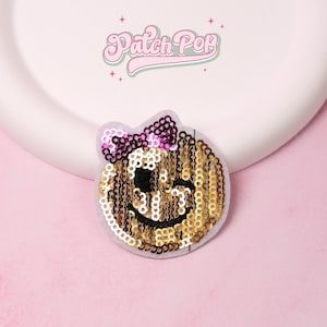 Può includere: Toppa con paillettes dorate a forma di faccina sorridente con un fiocco viola. La toppa ha dettagli neri e si trova su uno sfondo grigio. Il testo "Patch Pop" è sopra la toppa in rosa e verde.