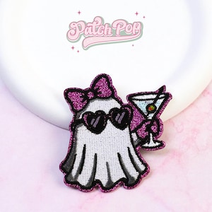 Può includere: Una toppa ricamata raffigurante un fantasma bianco con un fiocco viola glitterato e occhiali da sole neri a forma di cuore. Il fantasma tiene in mano un bicchiere da martini con un'oliva. La toppa ha un bordo viola glitterato e il testo Patch Pop.