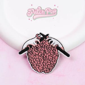 Puede incluir: Parche bordado que representa un cerebro rosa tejido con manos blancas y agujas negras. El parche tiene un borde blanco y el texto "Patch Pop" en rosa, en cursiva.