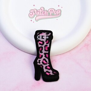 May include: An embroidered patch shaped like a knee-high platform boot with a black outline and a pink and white leopard print pattern. The boot features a black buckle detail at the top and a thick block heel. The text Patch Pop is visible above.