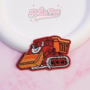May include: A red and orange embroidered patch featuring a cartoon bulldozer with a white outline. The patch has the text "Patch Pop" in a pink, cursive font.