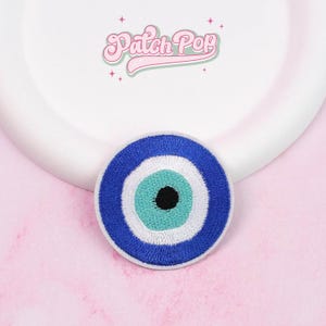 Puede incluir: Parche bordado redondo con un diseño de ojo turco azul y blanco y una pupila negra. El parche tiene un borde blanco. El texto "Patch Pop" está en escritura rosa encima del parche.