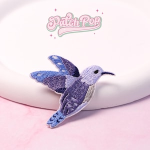 May include: Embroidered hummingbird patch in shades of blue and purple. The patch features detailed stitching, a long beak, and outstretched wings. The text "Patch Pop" is above the hummingbird in a retro font.