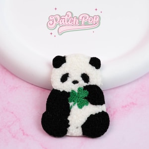 Puede incluir: Parche de oso panda negro, blanco y verde con un trébol de cuatro hojas. El parche tiene el texto "Patch Pop" en rosa. El fondo es rosa.