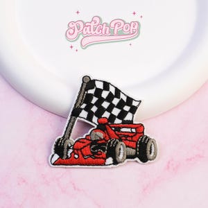 Pode incluir: Remendo bordado com um carro de corrida vermelho com pneus pretos e uma bandeira quadriculada. O remendo tem uma borda branca e o texto "Patch Pop" em rosa, em fonte cursiva.