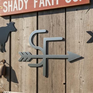 Op de afbeelding: Houten wanddecoratie met een metalen koe-silhouet, een bel en een metalen pijl met de letters "SF". Een rood bord bovenaan leest "SHADY FARM CO.". De decoratie is op een houten muur gemonteerd.