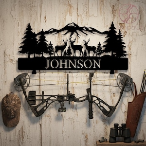 May include: Black metal wall art featuring a mountain range, trees, and deer silhouettes, with the name "JOHNSON" below. A compound bow is mounted below the art, with a camouflage cap, quiver, binoculars, and arrows. The wall is made of wood.