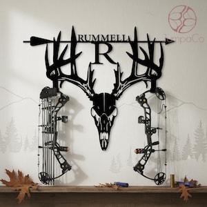 May include: Black metal wall art with a deer skull and antlers, the name "RUMMELL", and an arrow design. Two black compound bows are mounted below the skull. The wall art is displayed on a wooden shelf with autumn leaves and other items.