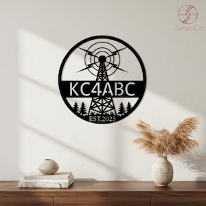 May include: Black metal wall art featuring a radio tower design within a circular frame. The text "KC4ABC" is prominently displayed, with "EST.2025" below. The artwork includes stylized trees and lightning bolts, suitable for home decor.