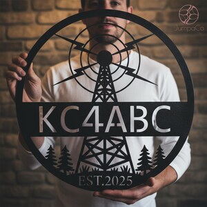 May include: Black metal wall art featuring a radio tower design with the call letters KC4ABC and the year EST.2025. The circular piece includes lightning bolts and stylized trees, suitable for home decor or a hobbyist's space.