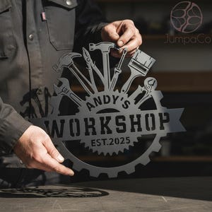 May include: A dark grey metal workshop sign featuring tools such as a hammer, spanner, and paintbrush. The sign is in the shape of a gear with the text "ANDY'S WORKSHOP EST. 2025". The sign is held by a person.