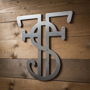 Op de afbeelding: Een metalen wandkunstwerk met de gestileerde letters "TF" en een dollarteken. Het kunstwerk is gemaakt van zilverkleurig metaal en gemonteerd op een achtergrond van houten planken. Het ontwerp is modern en geschikt voor woondecoratie.