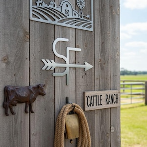 Op de afbeelding: Een rustieke houten muur versierd met metalen boerderijthema-decoraties. Inclusief een koeienbeeldje, een bord met de tekst "CATTLE RANCH", een bel met touw en een metalen pijl-vormig bord. Een metalen uitsnede van een boerderijsc&egrave;ne staat bovenaan.