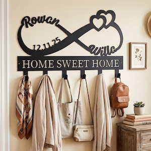 May include: Black metal wall-mounted coat rack with an infinity symbol design, featuring the names "Rowan" and "Willow" and the date "12.11.25". The rack includes hooks for hanging items, with the phrase "HOME SWEET HOME" below.