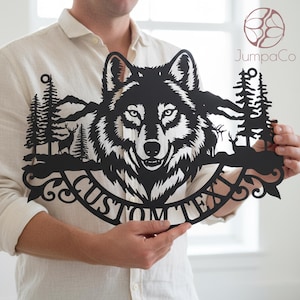 Op de afbeelding: Zwart metalen wandkunst met een wolfskop, bomen, bergen en herten. Het ontwerp bevat de tekst "CUSTOM TEXT" in een gebogen banner. Het kunstwerk wordt door een persoon vastgehouden.