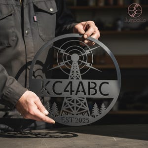 May include: A black metal sign featuring a radio tower design within a circular frame. The sign includes the text "KC4ABC" and "EST.2025". The design also incorporates stylized trees. The sign is held by a person.